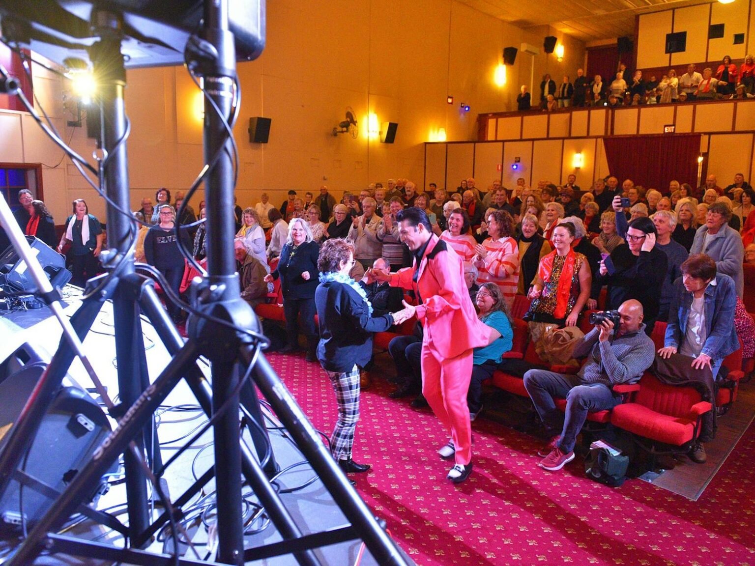 Elvis dances with audience member near stage as audience stands and joins in.