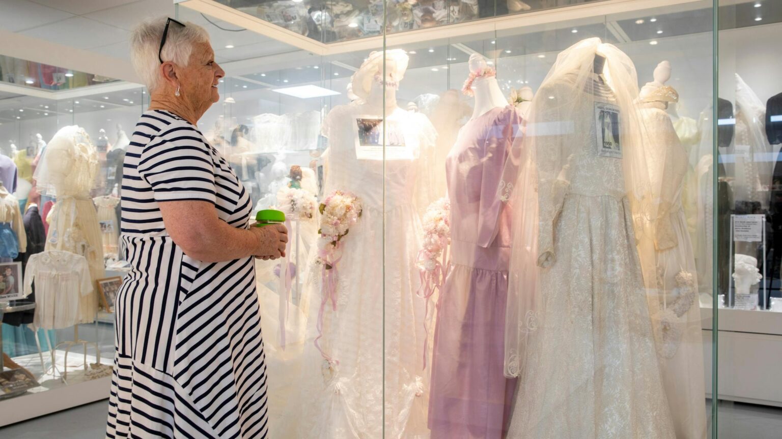 Shows a visitor viewing a selection of wedding dresses in glass cases in the clothing section.