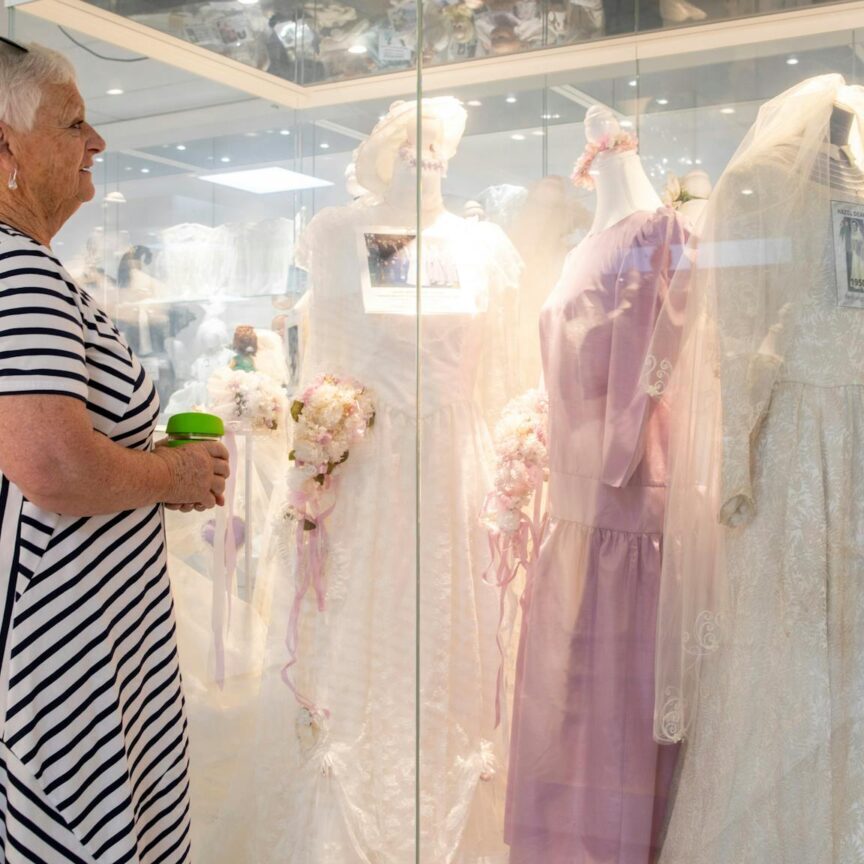 Shows a visitor viewing a selection of wedding dresses in glass cases in the clothing section.